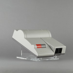 Op de afbeelding: Een grijs-witte videogameconsole met een rode en grijze knop. De console staat op een transparante acrylstandaard.