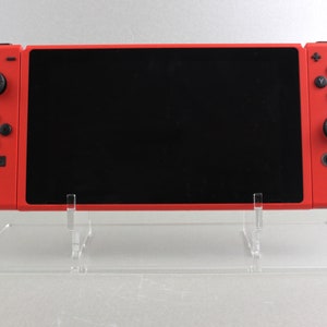 May include: A red handheld gaming console with a black screen and black buttons. The console is displayed on a clear acrylic stand. The stand has a rectangular base and two vertical supports.