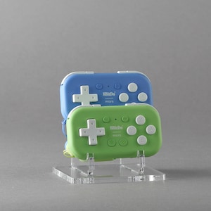 Könnte beinhalten: Zwei Handheld-Gamecontroller, einer blau und einer grün, werden auf einem transparenten Acrylständer präsentiert. Jeder Controller hat ein weißes Steuerkreuz und weiße Tasten. Der Text "8BitDo micro" ist aufgedruckt.