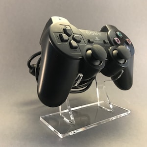 Könnte beinhalten: Ein schwarzer PlayStation 2-Controller mit einem transparenten Acryl-Ständer. Der Controller trägt das Sony PlayStation-Logo und die Aufschrift "PlayStation 2".