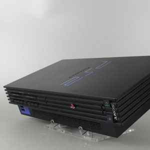 Può includere: Una console PlayStation 2 nera con il logo PlayStation sulla parte superiore. La console è posizionata su un supporto in acrilico trasparente.