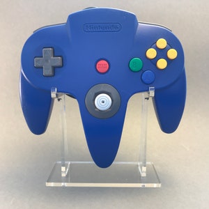 Könnte beinhalten: Ein blauer Nintendo 64-Controller auf einem transparenten Acryl-Ständer. Der Controller hat eine graue Frontplatte mit einem weißen Analogstick und einem roten Startknopf. Der Controller hat gelbe, grüne und blaue Knöpfe.