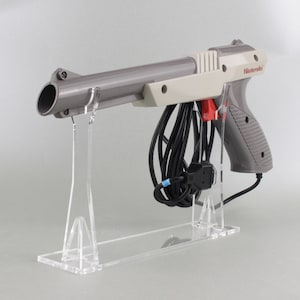 Könnte beinhalten: Eine graue und weiße Nintendo Zapper Lichtpistole für die NES-Konsole, mit einem schwarzen Kabel und einem durchsichtigen Kunststoffständer.