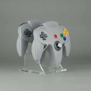 May include: Two gray Nintendo 64 controllers on a clear acrylic stand. The controllers have a gray and black color scheme with colored buttons.