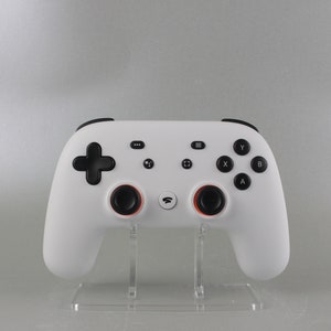 May include: A white games controller with black buttons and a black directional pad. The controller has two black analogue sticks with orange accents and is displayed on a clear acrylic stand. The controller has the Stadia logo.
