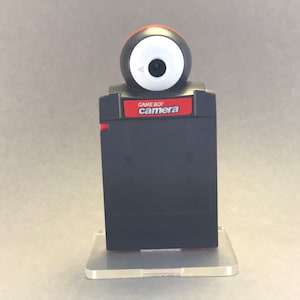 Può includere: Una cartuccia nera di Game Boy Camera con un obiettivo rosso e bianco in cima. La cartuccia ha il testo "GAME BOY camera" stampato su di essa.
