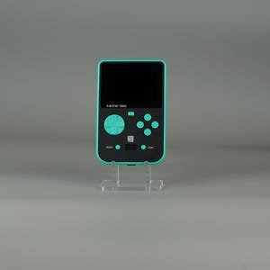 Può includere: Una console per videogiochi portatile di colore verde acqua e nero con un pad direzionale e quattro pulsanti. La console ha un piccolo schermo e il testo "TAITO 1983" stampato sul davanti.