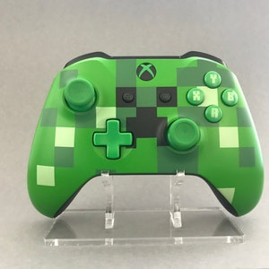 May include: Green and black Xbox controller with a pixelated design. The controller has green buttons and joysticks, with the Xbox logo at the top. The design resembles a character from the game Minecraft. The controller is displayed on a clear acrylic stand.