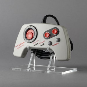May include: A gray NES Advantage joystick with red buttons and a black cord. The joystick is on a clear acrylic stand. The text "NES Advantage" and "Nintendo" is printed on the joystick.