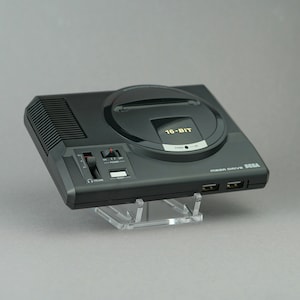 May include: A black Sega Mega Drive 16-bit video game console with a grey and black controller and a black cartridge slot. The console is sitting on a clear acrylic stand.