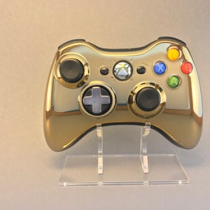 May include: A gold-coloured Xbox 360 controller with black buttons and a clear acrylic stand. The controller has a glossy finish and the buttons are labelled with the letters X, Y, A, and B.