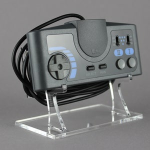 Könnte beinhalten: Eine schwarze Sega Genesis Konsole mit einem grauen Controller und einem schwarzen Kabel. Die Konsole steht auf einem durchsichtigen Acryl-Ständer.