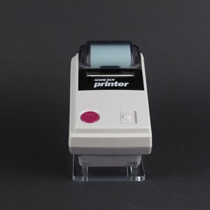 May include: A white Game Boy Printer with a black and white display and a pink button. The printer is on a clear stand.
