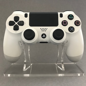 May include: A white PlayStation 4 controller is displayed on a clear acrylic stand. The controller has black buttons and a black touchpad.
