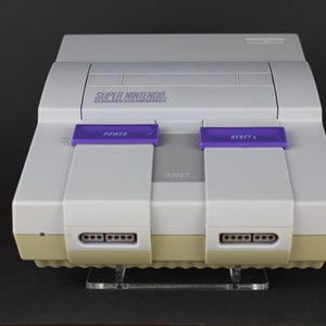 Peut inclure: Une console Super Nintendo Entertainment System blanche avec des boutons violets indiquant "POWER" et "RESET". La console a un fond beige avec deux rangées de ports de connexion.