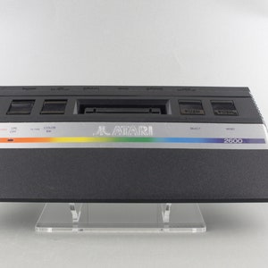 Könnte beinhalten: Eine schwarze Atari 2600 Videospielkonsole mit einer silbernen Frontplatte. Die Konsole hat einen Steckplatz für eine Spielkassette, zwei Controller-Anschlüsse und einen Netzschalter. Die Frontplatte trägt das Atari-Logo und den Text "2600".