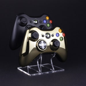 May include: Two video game controllers, one black and one gold, are displayed on a clear acrylic stand. The controllers have coloured buttons and a black and white directional pad.