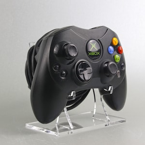 Könnte beinhalten: Ein schwarzer Xbox-Controller mit grünen und weißen Knöpfen auf einem transparenten Acryl-Ständer. Der Controller hat ein schwarzes Kabel.