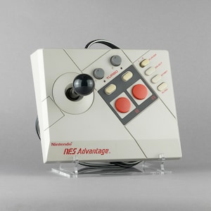 Może przedstawiać: Biało-szary joystick Nintendo NES Advantage z czarną gałką joysticka i czerwonymi przyciskami. Joystick ma przycisk turbo i wiele innych przycisków, w tym select, start i reset. Joystick znajduje się na przezroczystym akrylowym stojaku.