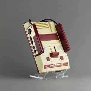 Peut inclure: Une console de jeu vidéo Nintendo Entertainment System (NES) vintage de couleur rouge et blanche. La console est sur un support en acrylique transparent et a une manette attachée à l'avant. La console porte l'inscription "FAMILY COMPUTER" sur le devant.