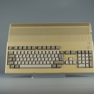 Könnte beinhalten: Ein beigefarbener Amiga-Computer mit einer vollständigen Tastatur. Die Tastatur hat hellgraue Tasten und einen Ziffernblock. Das Wort "AMIGA" ist oben auf dem Computer aufgedruckt. Der Computer steht auf einem transparenten Ständer.