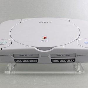 Könnte beinhalten: Weiße PSone-Konsole mit Sony-Logo und dem Text "PSone". Die Konsole verfügt über ein Laufwerk, Ein-/Aus- und Reset-Tasten sowie zwei Speicherkartensteckplätze. Präsentiert auf einem transparenten Acryl-Ständer.