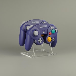 May include: Two purple Nintendo GameCube controllers on a clear acrylic stand. The controllers have various colored buttons, including green, red, and yellow. The GameCube logo is visible on the top of the controllers. The stand is square-shaped.