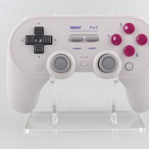 Könnte beinhalten: Ein weißer 8BitDo Pro 2 Gaming-Controller mit grauen Tasten und rosafarbenen Fronttasten. Der Controller steht auf einem durchsichtigen Plastikständer.