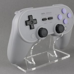 May include: A gray and black video game controller with purple buttons, sitting on a clear acrylic stand. The controller has the text "8BitDo SN30 Pro+" printed on it.