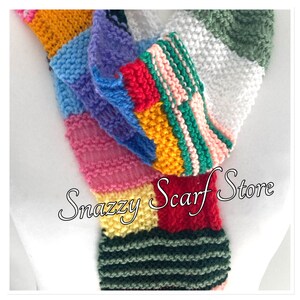 May include: A colourful knitted scarf with a patchwork design in shades of pink, red, yellow, green, blue, and white. The scarf is folded in half and the text "Snazzy Scarf Store" is visible in the lower right corner.
