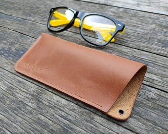 May include: A brown leather glasses case with the word "Criggan" embossed on the front. The case is closed and lying on a wooden surface. A pair of yellow-tinted glasses with black frames is lying next to the case.