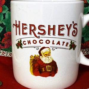 May include: White ceramic mug featuring the Hershey's Chocolate logo in red lettering with a Santa Claus illustration. The Santa illustration shows Santa with a white beard and a red suit, carrying a bag of gifts. The mug has a white handle.