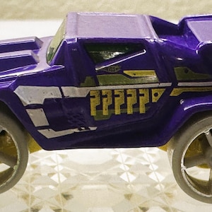 May include: A purple toy car with a rugged, angular body design, featuring large light gray wheels, a rear spoiler, and yellow accents on the side panels. The vehicle has a low-profile windshield and a distinct, boxy frame with white and yellow graphic details along the doors.