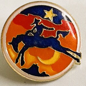 May include: A round pin with a cowboy riding a bucking bronco. The pin features a blue cowboy and horse against a sunset background of orange, red, and yellow. A yellow star and crescent moon are also present.