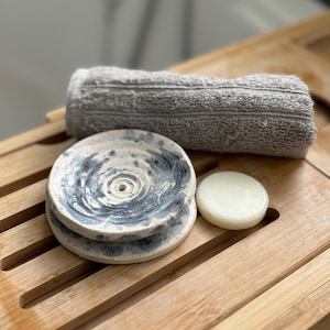 Op de afbeelding: Een witte keramische zeepbak met een blauw swirl-ontwerp. De bak is gestapeld op een andere bak. Een witte zeepreep ligt op een houten oppervlak naast de bak.