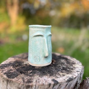 Può includere: Una tazza in ceramica blu chiaro con un viso scolpito sul davanti. La tazza è appoggiata su un ceppo d'albero.