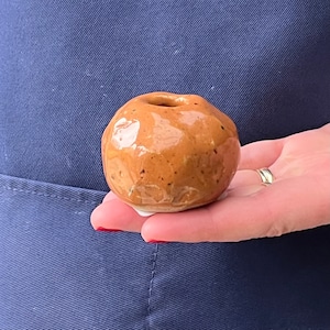 May include: A small, round, brown ceramic pot with a smooth, glazed surface. The pot has a small opening at the top.
