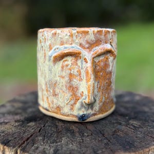 May include: A handmade ceramic mug with a face design. The mug has a textured surface with a mix of earthy colours, including brown, tan, and touches of blue. The face is sculpted with defined features, creating a unique and artistic piece.