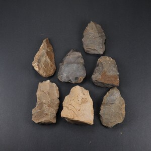 May include: Seven hand-shaped, gray and tan stone tools are arranged on a dark gray surface. The tools vary in size and shape, with sharp edges and rough textures, suggesting they are ancient artifacts.