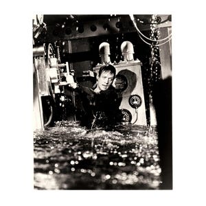 May include: A black and white photograph showing a person submerged in water inside a room with industrial machinery. The person is reaching toward a large circular valve on a wall panel featuring multiple round gauges and pipes.