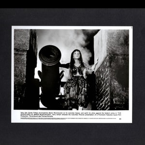 May include: A black and white photo of a young girl standing in front of a cannon. She is wearing a long, flowing dress and has her hair down. The photo is from the movie "The Adventures of Baron Munchausen".