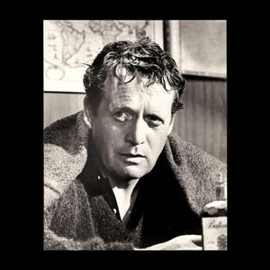 May include: A black and white portrait of a person with wavy hair, wearing a dark collared shirt under a thick, textured wool blanket. The person has a focused expression, looking directly forward. A bottle with a partial label is visible in the foreground.