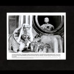 May include: A black and white photo of a woman in a white costume with a crown and a white feathered collar. She is sitting on a bed and tickling her feet. The photo is from the movie "The Adventures of Baron Munchausen".