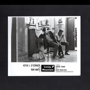 May include: Black and white photo of two men sitting on a bench in front of a piano. The men are wearing casual clothing and one is holding a cigarette. The photo is from the movie "Candy Mountain" starring Kevin J. O'Connor and Tom Waits.