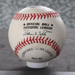 May include: A baseball with red stitching and the text "OFFICIAL BALL NATIONAL LEAGUE" and the signature "William D. White PRES". The ball also has the text "CUSHIONED CORK CENTER" and "RO-N".
