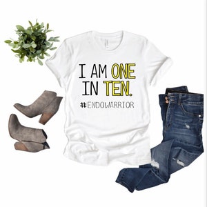 May include: White t-shirt with black text that reads "I am one in ten. #EndoWarrior". The shirt is on a white background with a pair of gray ankle boots and blue jeans.