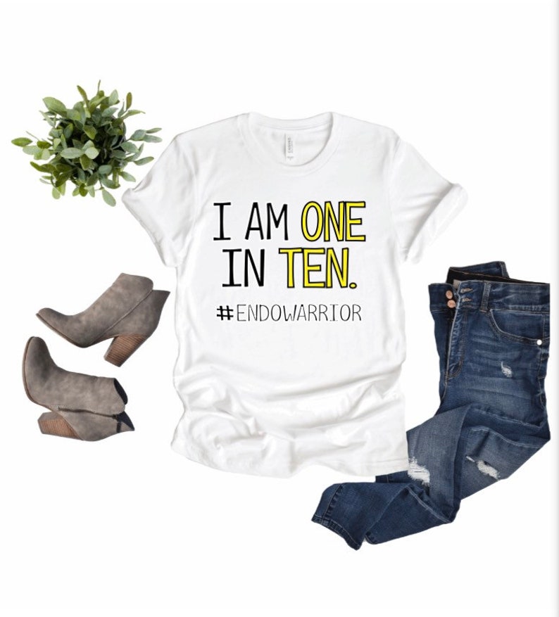 May include: White t-shirt with black text that reads "I am one in ten. #EndoWarrior". The shirt is on a white background with a pair of gray ankle boots and blue jeans.
