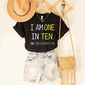 May include: Black t-shirt with the text "I am one in ten. #Endowarrior" in yellow lettering. The shirt is on a hanger with a straw hat and a straw bag.