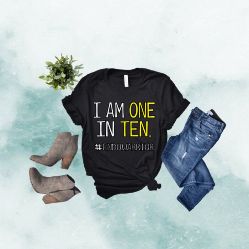 May include: Black t-shirt with the text "I AM ONE IN TEN." in white with a yellow outline. Below the text is the hashtag "#ENDOWARRIOR" in yellow. The shirt is laying on a light blue background with a pair of jeans and brown ankle boots.