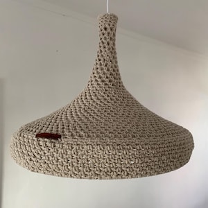 Puede incluir: Una lámpara colgante de crochet beige con una pantalla ancha y redonda. La pantalla tiene una textura tejida y una pequeña etiqueta de cuero marrón.